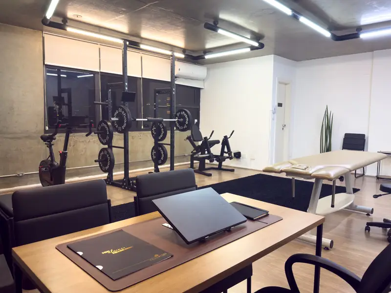 Área de consulta da Joelho Mais Fisio com mesa, notebook e maca, ao lado dos equipamentos de treino