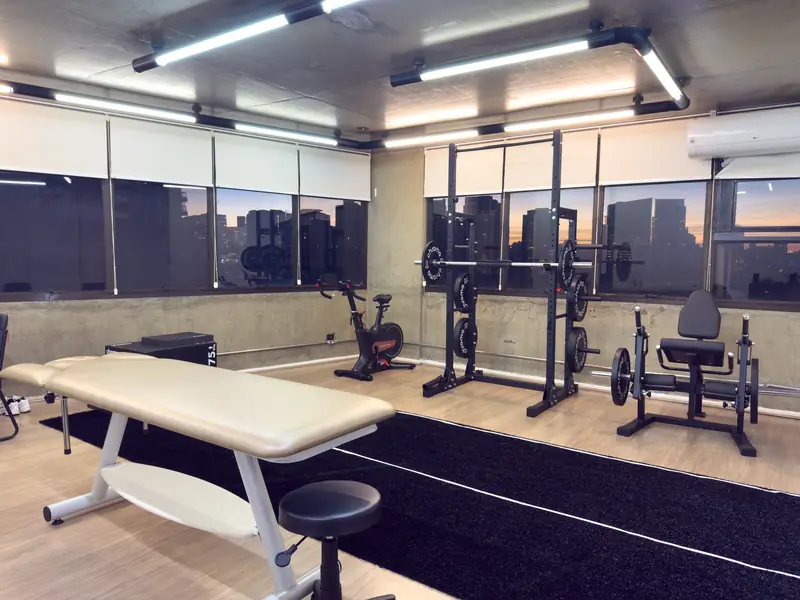 Sala de reabilitação da Joelho Mais Fisio com maca, rack de agachamento, bicicleta ergométrica e cadeira extensora, com vista para a cidade